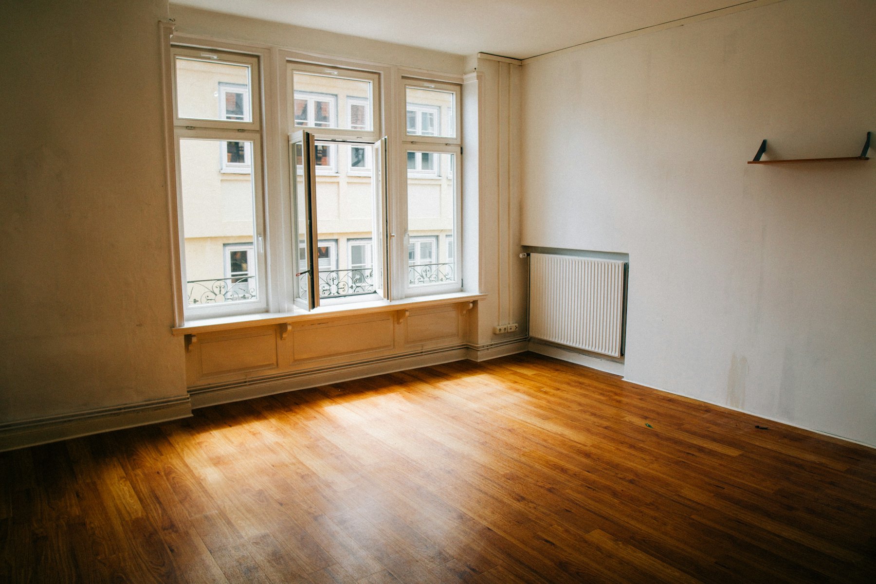 Ein leeres Altbauzimmer mit großem, geöffnetem Fenster; Licht liegt auf dem Holzboden.